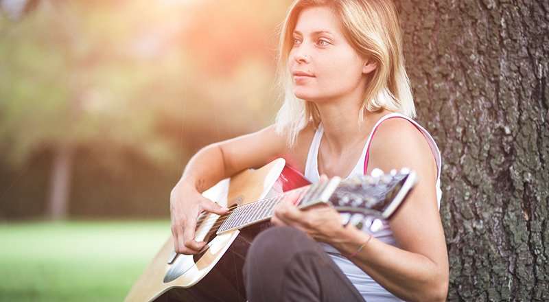 Junge Frau, die im Grünen an einen Baum gelehnt, Gitarre spielt.